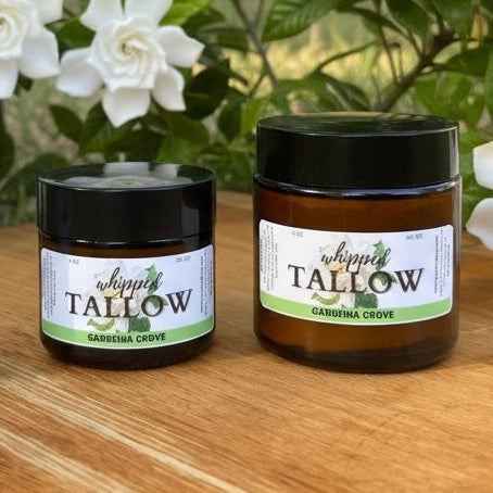 Two jars of 'Whipped Tallow' with a gardenia grove scent on a wooden surface with flowers in the background.