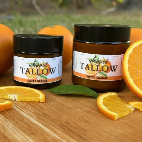 Two jars of 'Whipped Tallow Sweet Orange' on a wooden surface with oranges and leaves.